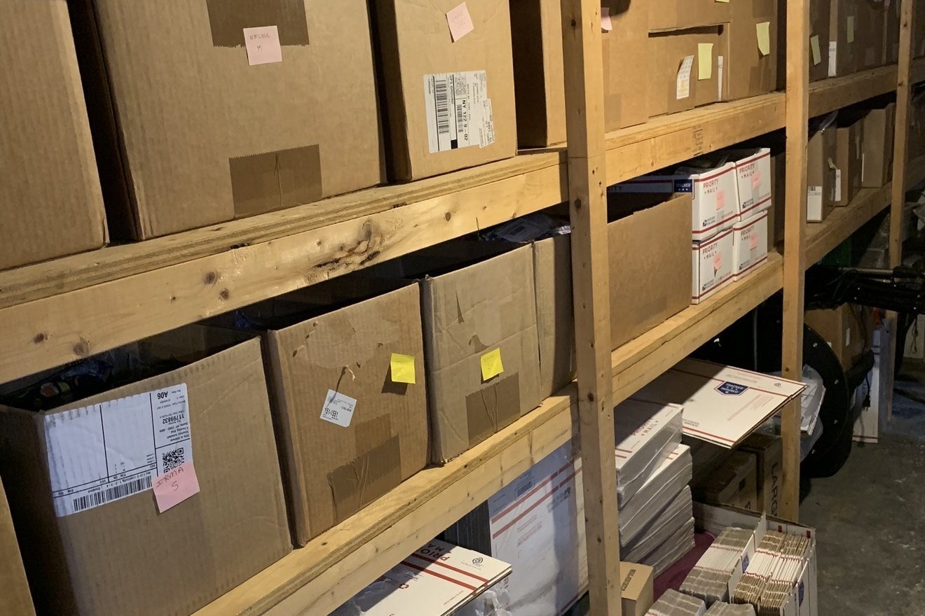 A basement shipping area with cartons, shelving and stacks of Priority Mail boxes.