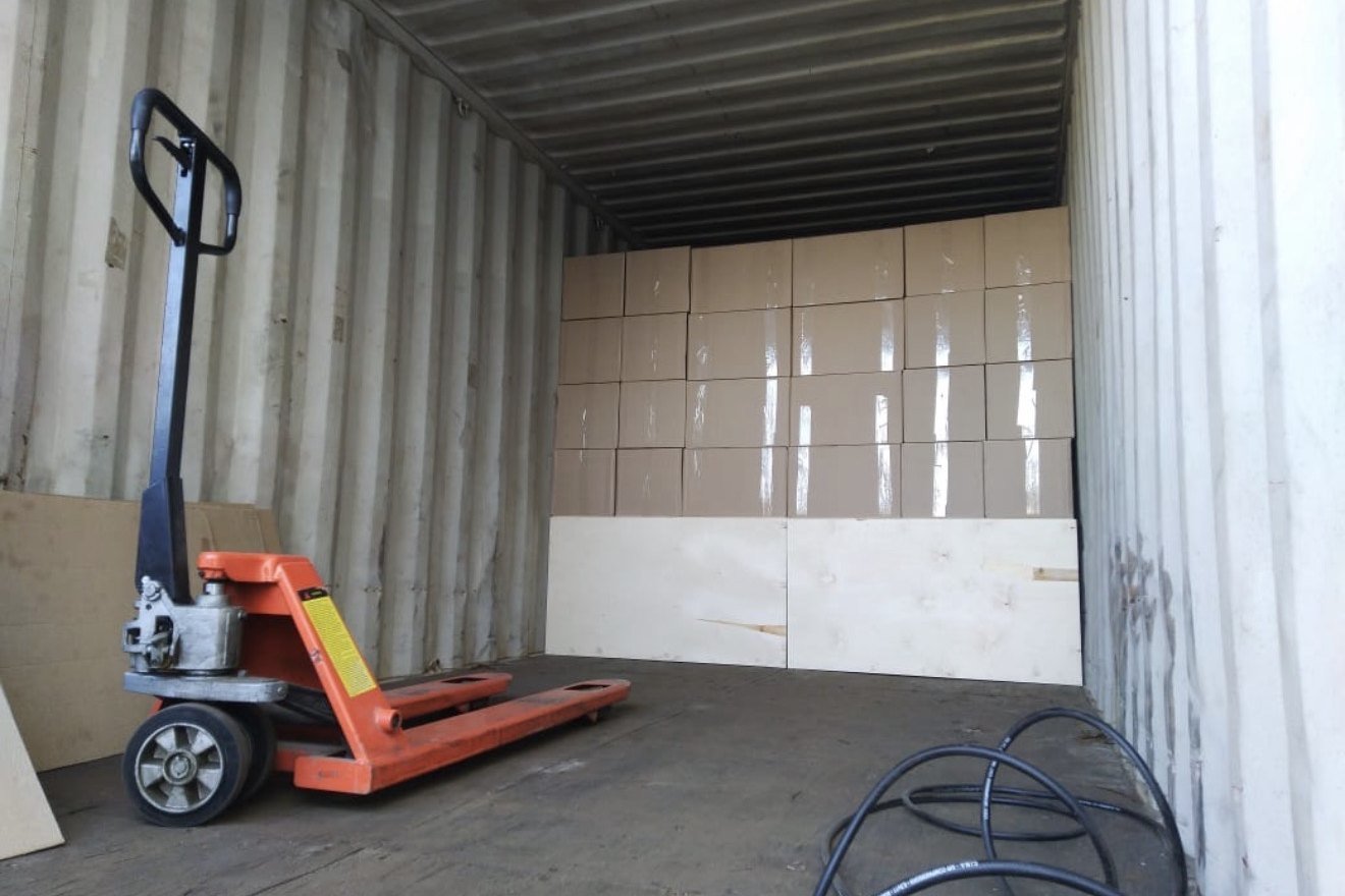Inside a freight container with boxed inventory stacked at the far end and a pallet jack in the foreground.